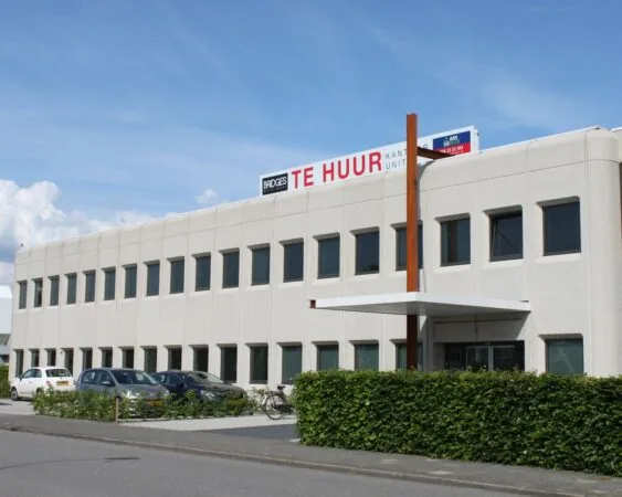 Kantoorgebouw met een 'Te Huur' bord op het dak, gelegen aan een straat met geparkeerde auto's en fietsen.