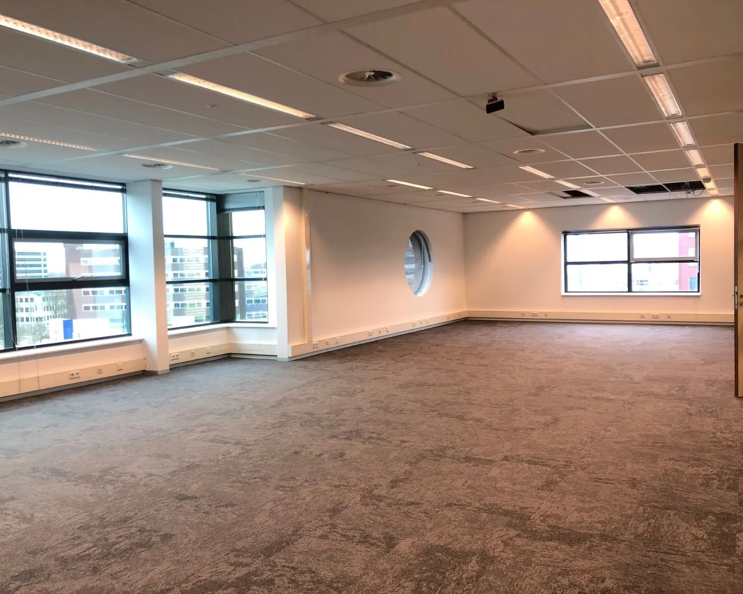 Leeg kantoorpand met grijze vloerbedekking, systeemplafond en grote ramen met uitzicht op een stedelijke omgeving.