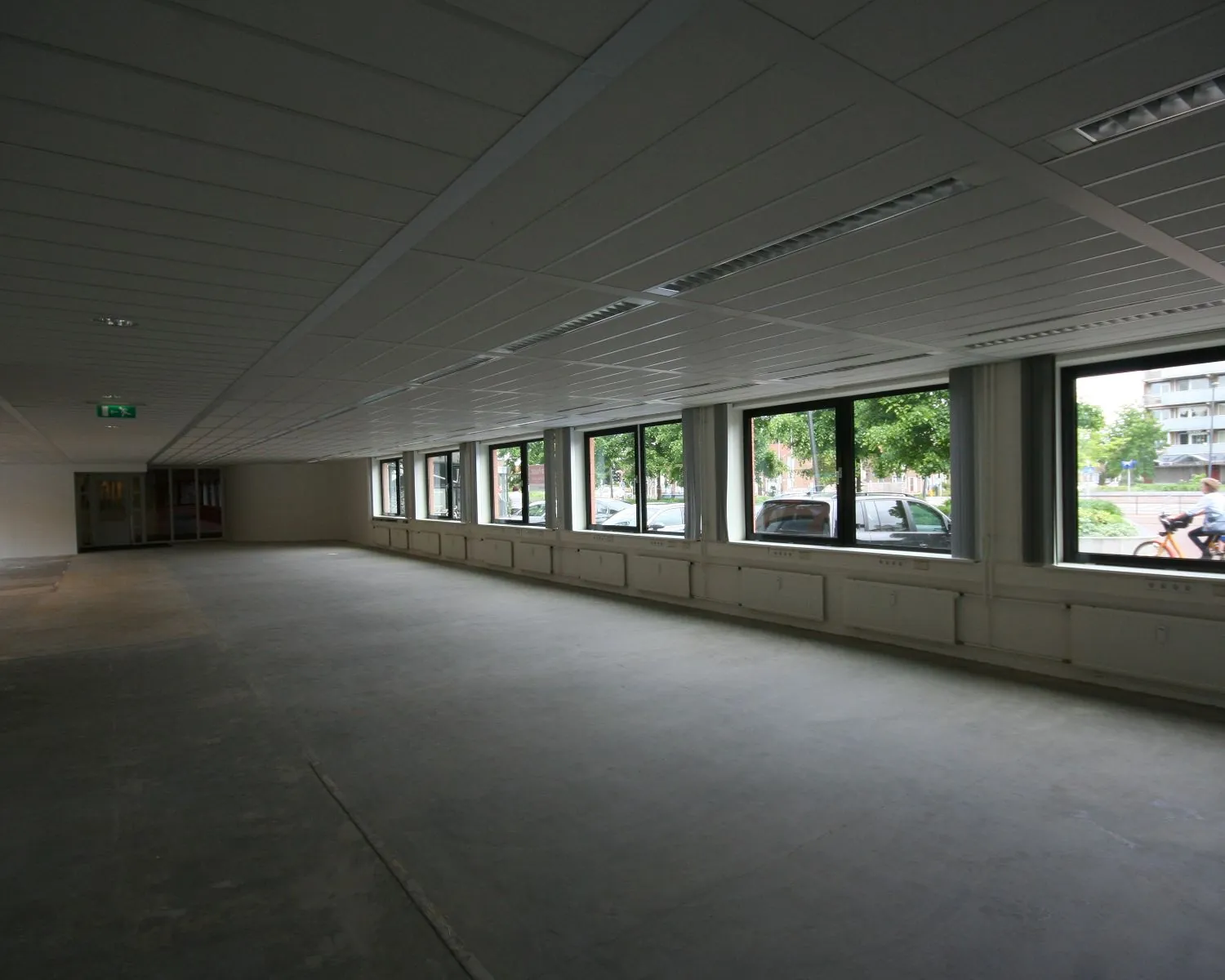 Lege kantoorruimte met systeemplafond, grote ramen en uitzicht op een straat met geparkeerde auto's.
