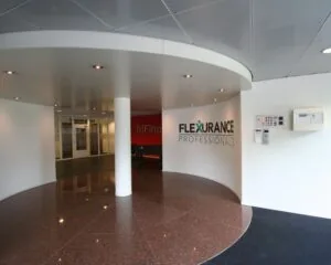 Ingang van een modern kantoorgebouw met de logo’s van Flexurance Professionals en TriFina aan de muur.