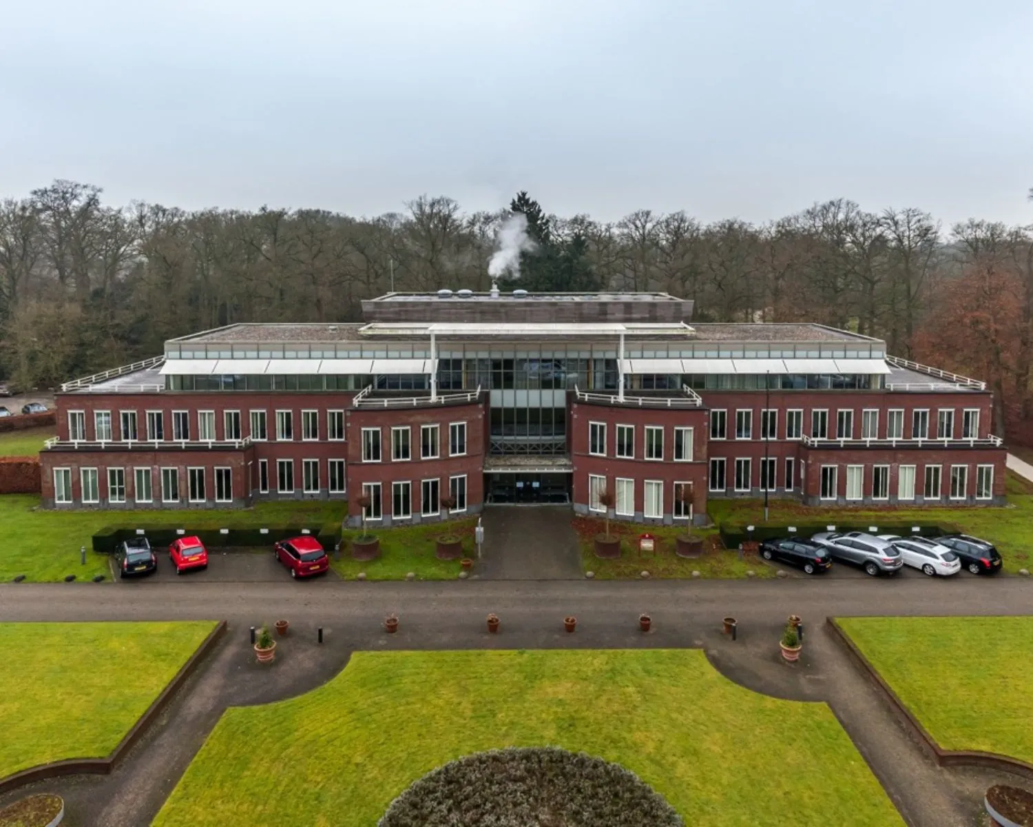 Luchtfoto van een modern kantoorgebouw met rode bakstenen gevel en omliggende parkeerplaatsen, omgeven door een bosrijke omgeving.