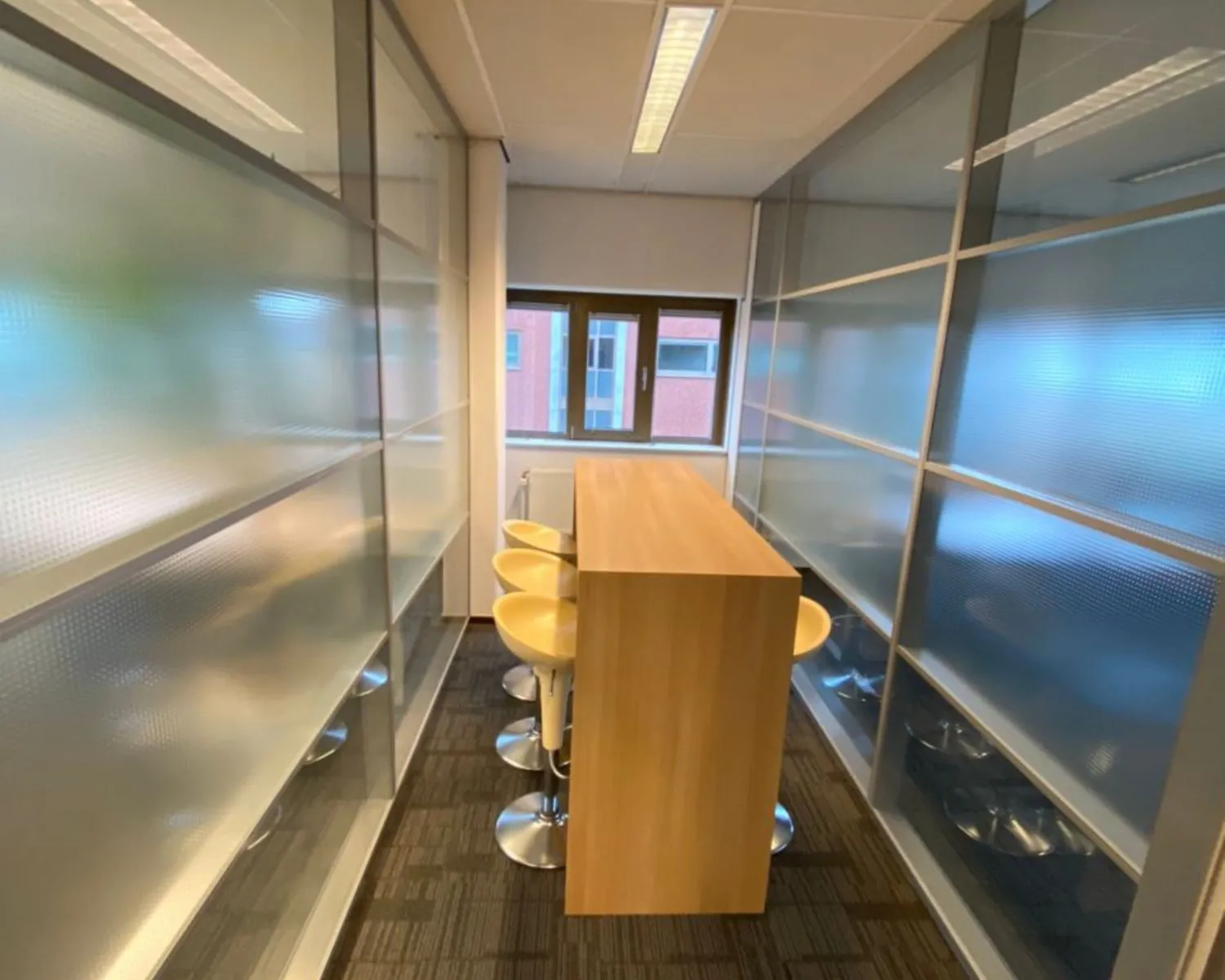 Smalle vergaderruimte met hoge houten tafel en gele barkrukken tussen matglazen wanden.