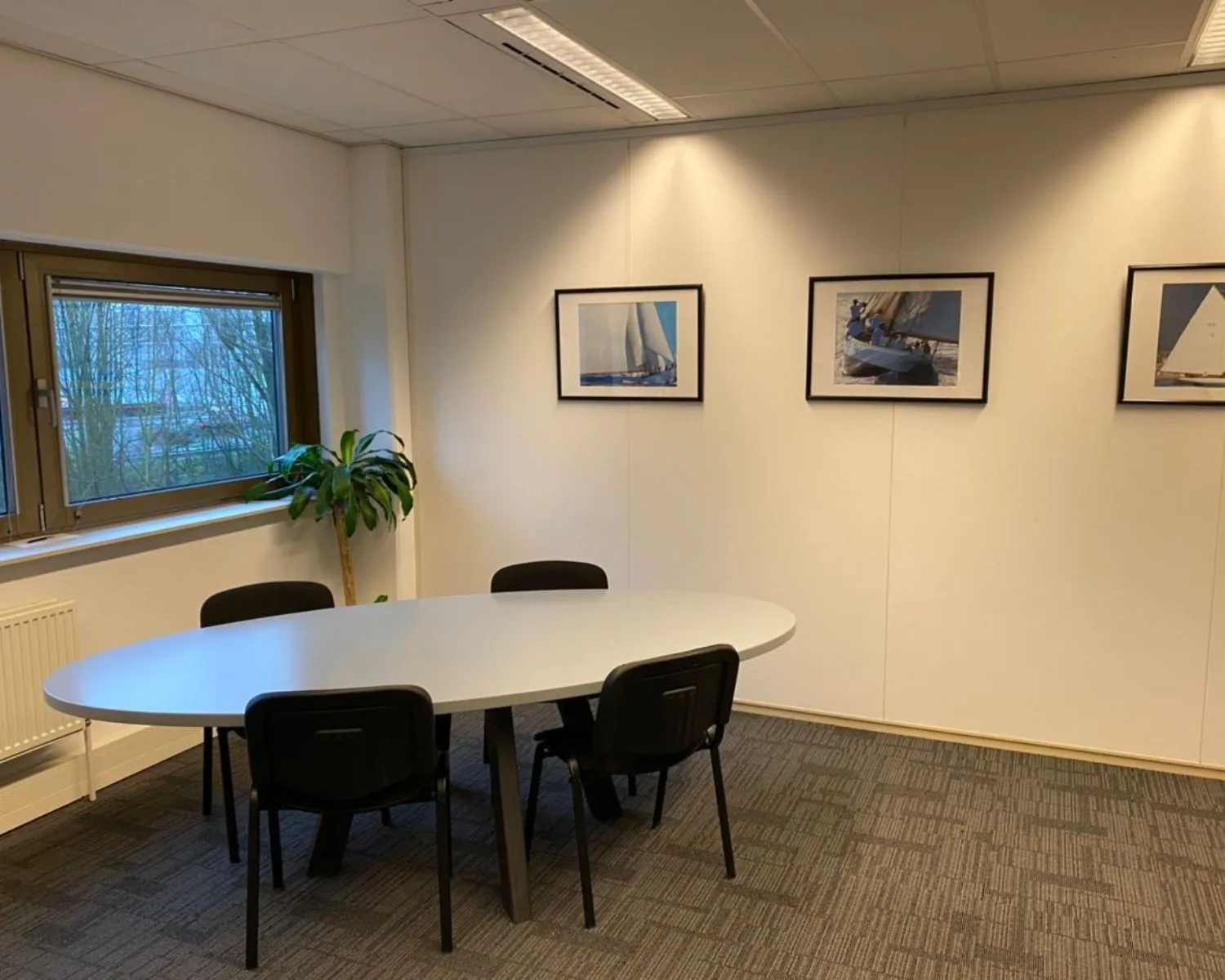 Kleine vergaderruimte met ovale tafel, vijf stoelen, een plant bij het raam en drie foto's van zeilboten aan de muur.