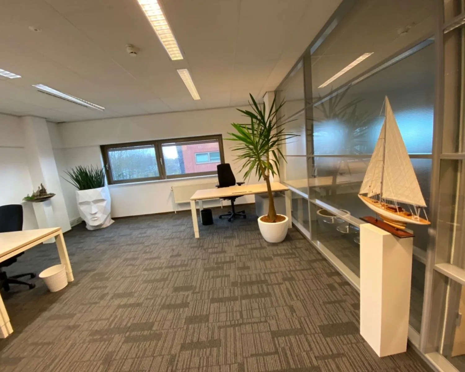 Modern kantoor met bureaus, planten, een groot modelzeilschip en decoratief hoofdbeeld.