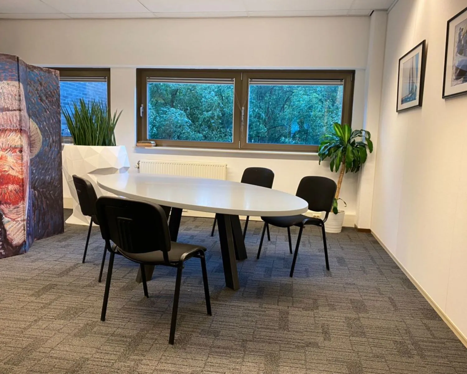 Vergaderruimte met ovale tafel, zes zwarte stoelen, grote ramen, planten en een kamerscherm met een schildering van Van Gogh.