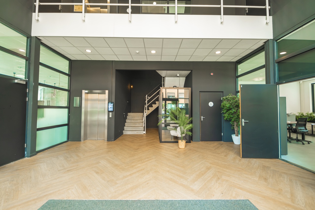 Ingang van een modern kantoorpand met een lift, trap, planten en glazen kantoorruimtes.