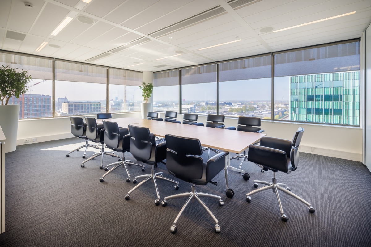 Een moderne vergaderruimte met grote ramen, een lange tafel en zwarte stoelen.