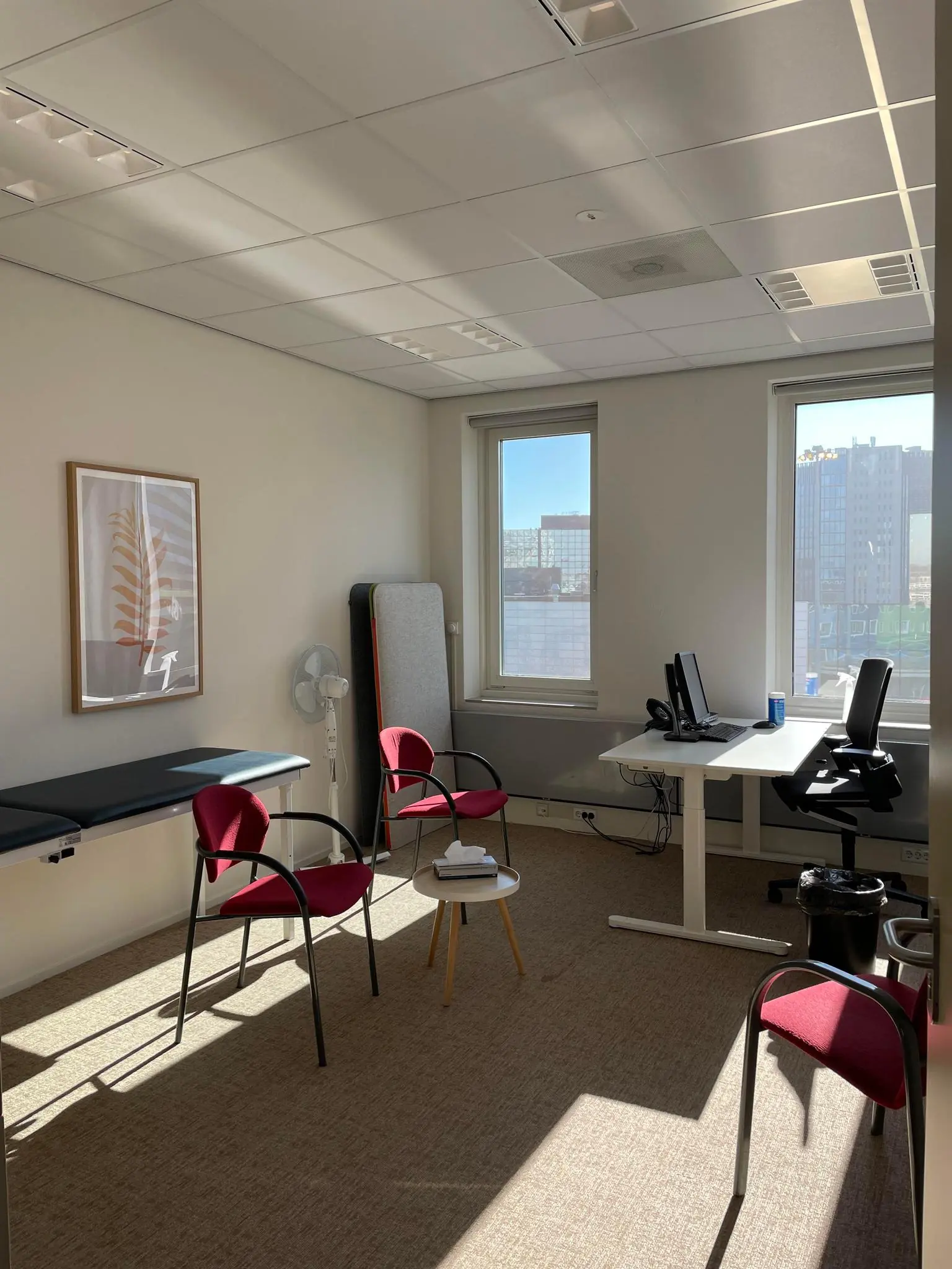 Licht en modern ingericht spreek- of dokterskantoor aan de Johan Cruijff Boulevard met bureau, stoelen en onderzoeksbank.