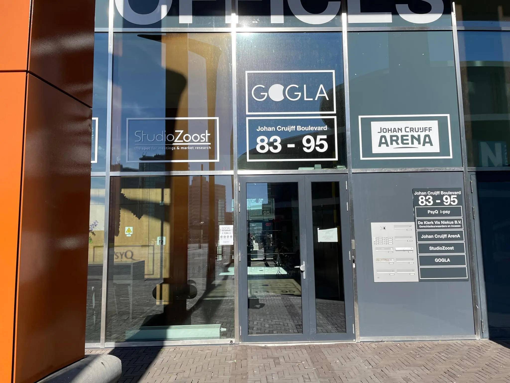 Ingang van een kantoorgebouw aan de Johan Cruijff Boulevard 83-95 met borden van StudioZoost, GOGLA en Johan Cruijff Arena.