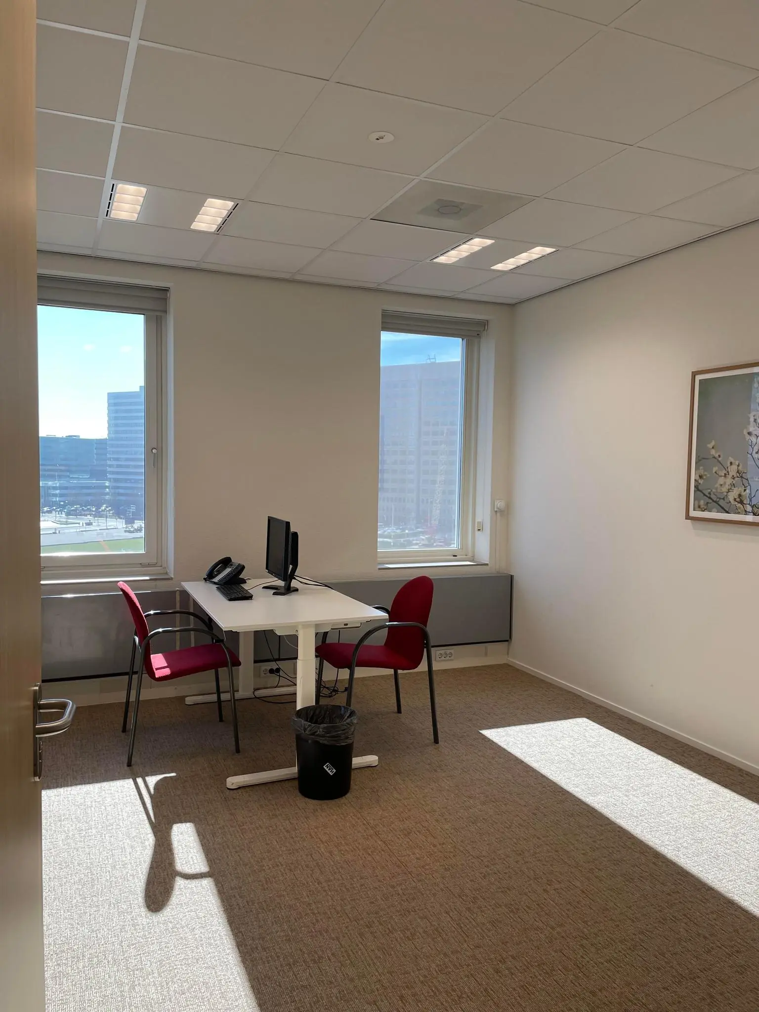 Klein en licht kantoor met twee rode stoelen, een bureau met computer en uitzicht op kantoorgebouwen aan de Johan Cruijff Boulevard.