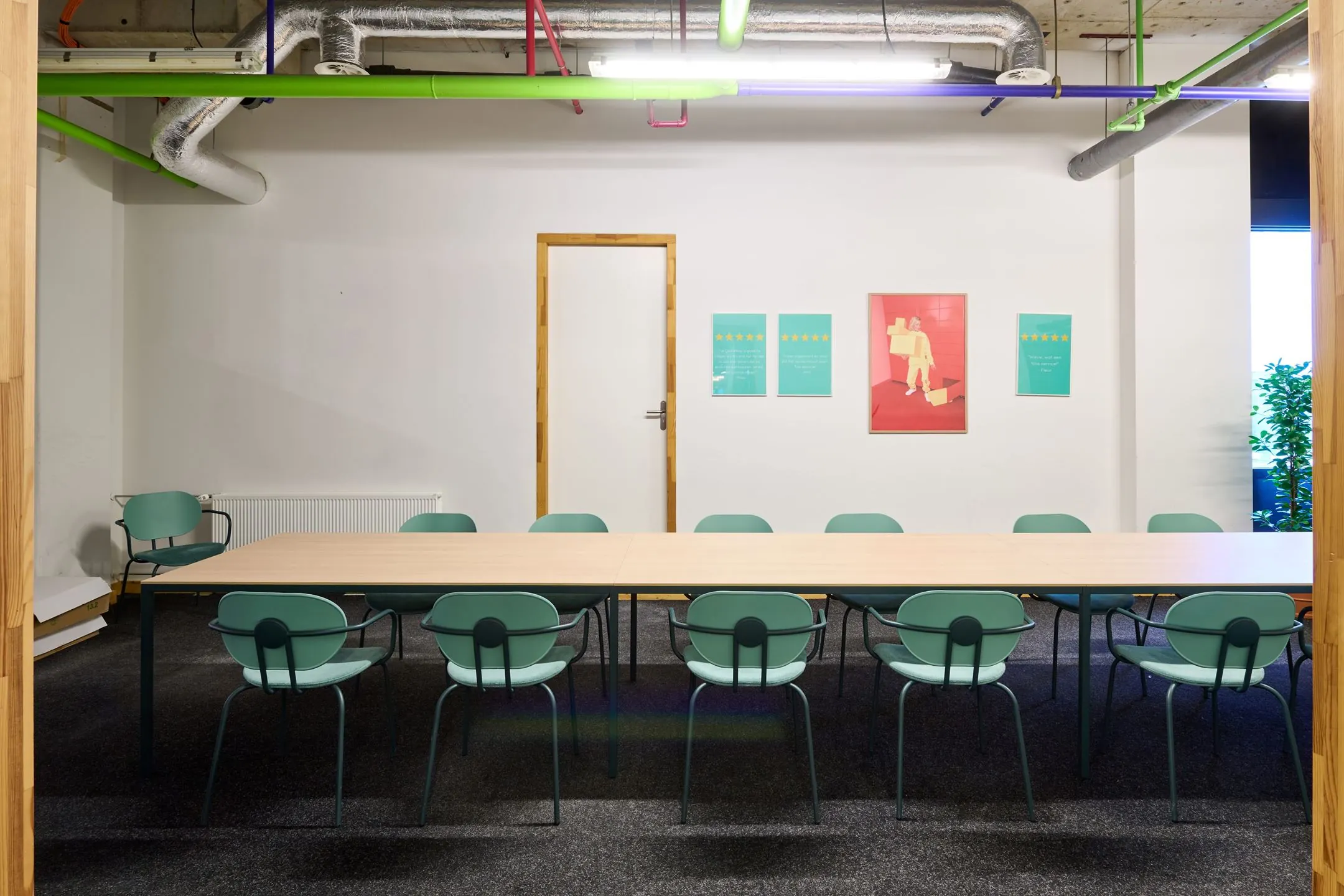 Vergaderruimte met lange tafel, groene stoelen en felgekleurde buizen aan het plafond op Johan Huizingalaan 763a.