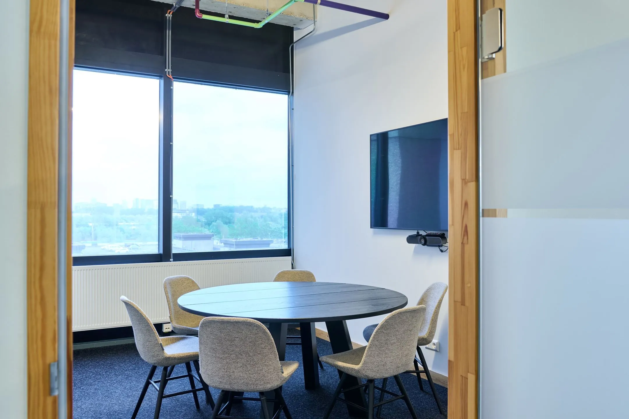 Kleine vergaderruimte met ronde tafel, zes stoelen, groot raam en scherm aan de muur op Johan Huizingalaan 763a.