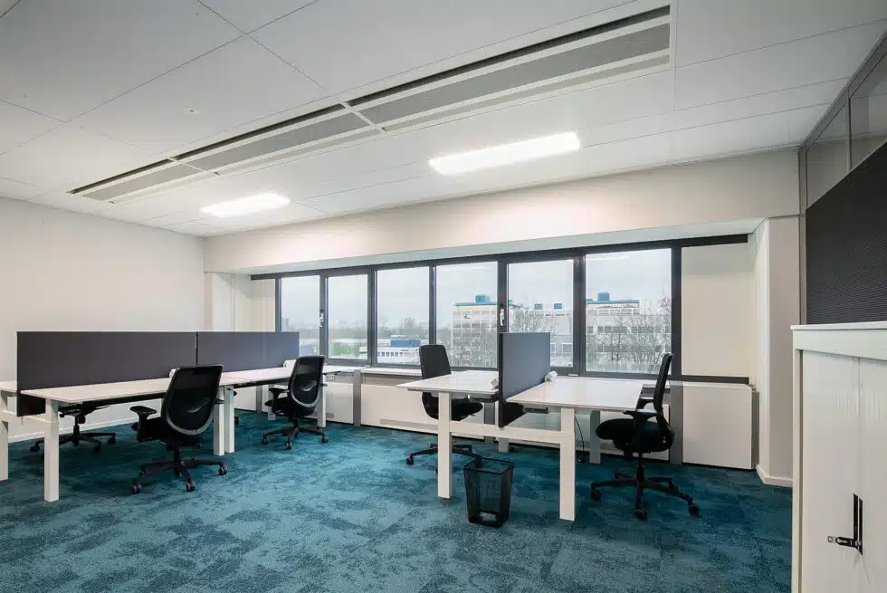 Een modern kantoor met vier lege bureaus en zwarte stoelen bij grote ramen.