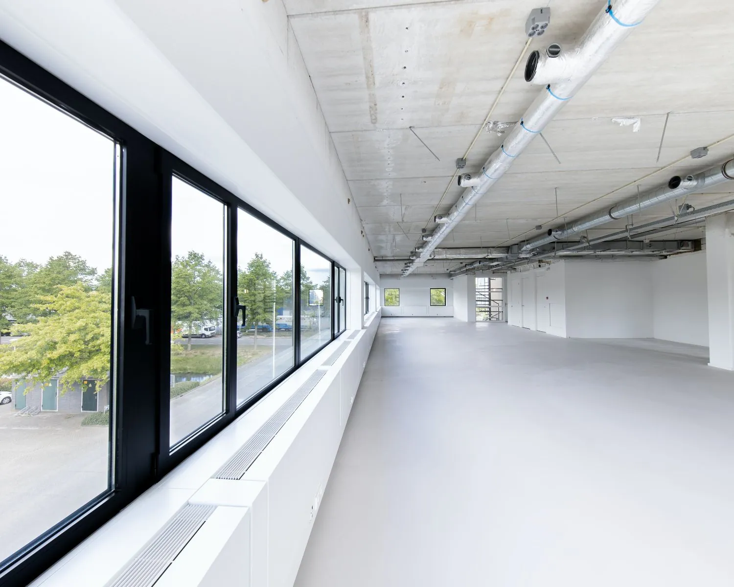 Lichte, moderne kantoorruimte te huur aan de Basicweg 12 in Amersfoort met grote ramen en een betonnen plafond met zichtbare leidingen.