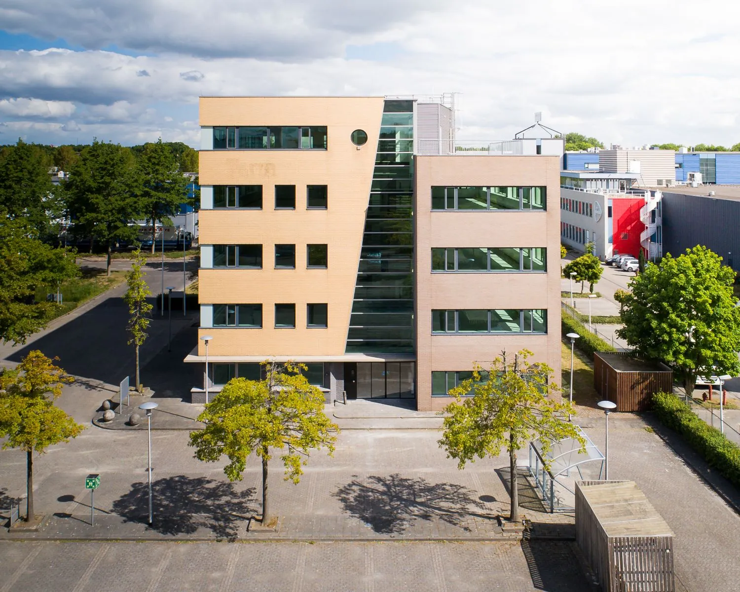Modern kantoorgebouw aan de Basicweg 12 in Amersfoort, met glazen centrale gevel en omringd door bomen en parkeerplaatsen.