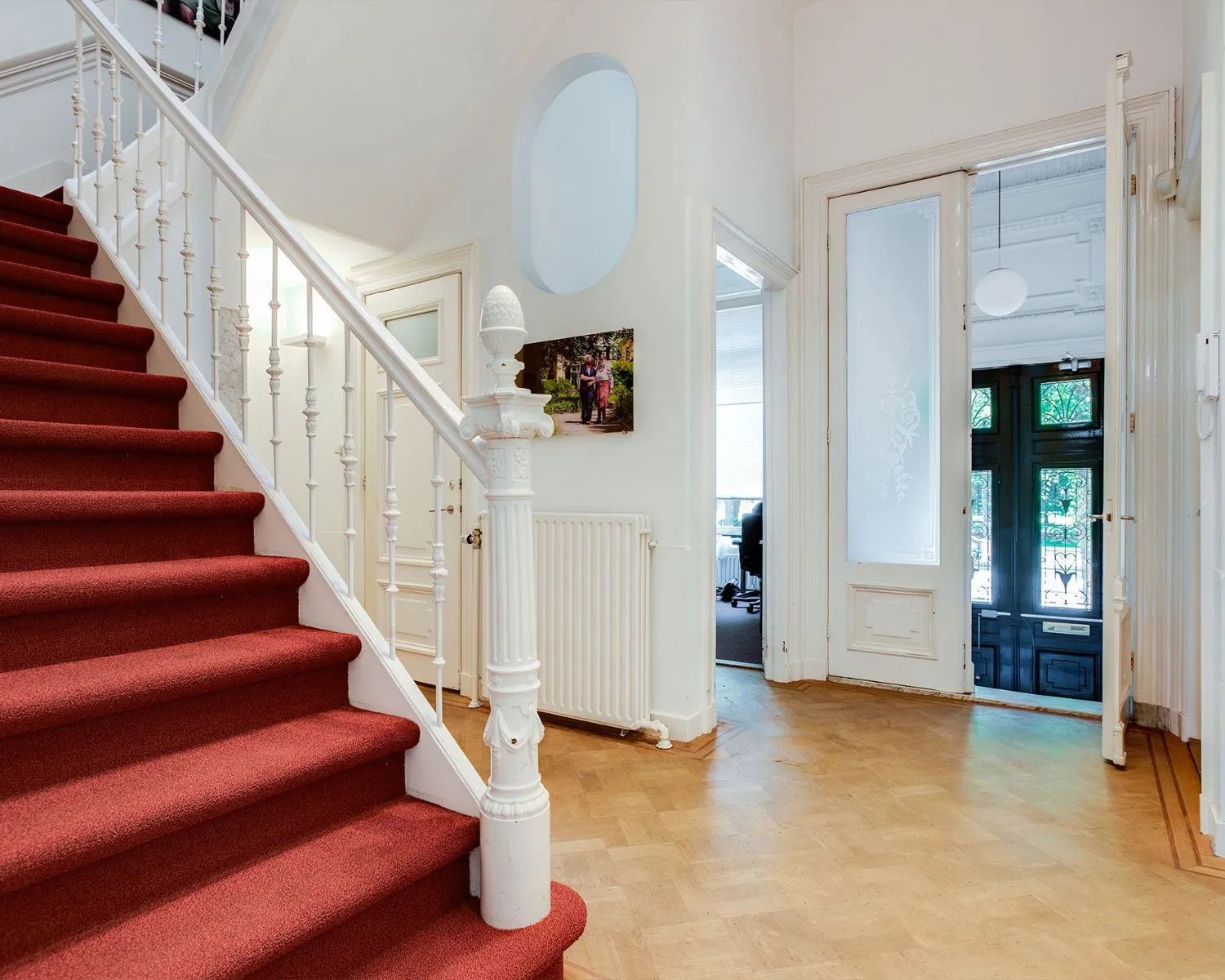 Entreehal van een kantoorruimte met houten vloer, rode trapbekleding en zicht op een kantoorruimte en voordeur aan de Dreef 36 in Haarlem.