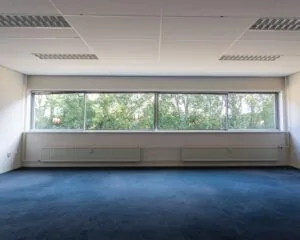 Lege kantoorruimte met blauwtapijt en grote ramen aan de Fruitweg 36-60, met uitzicht op groene bomen.