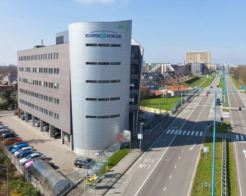 Modern kantoorgebouw aan de Groeneweg 2-1 met parkeerplaats en zicht op een brede stadsweg.