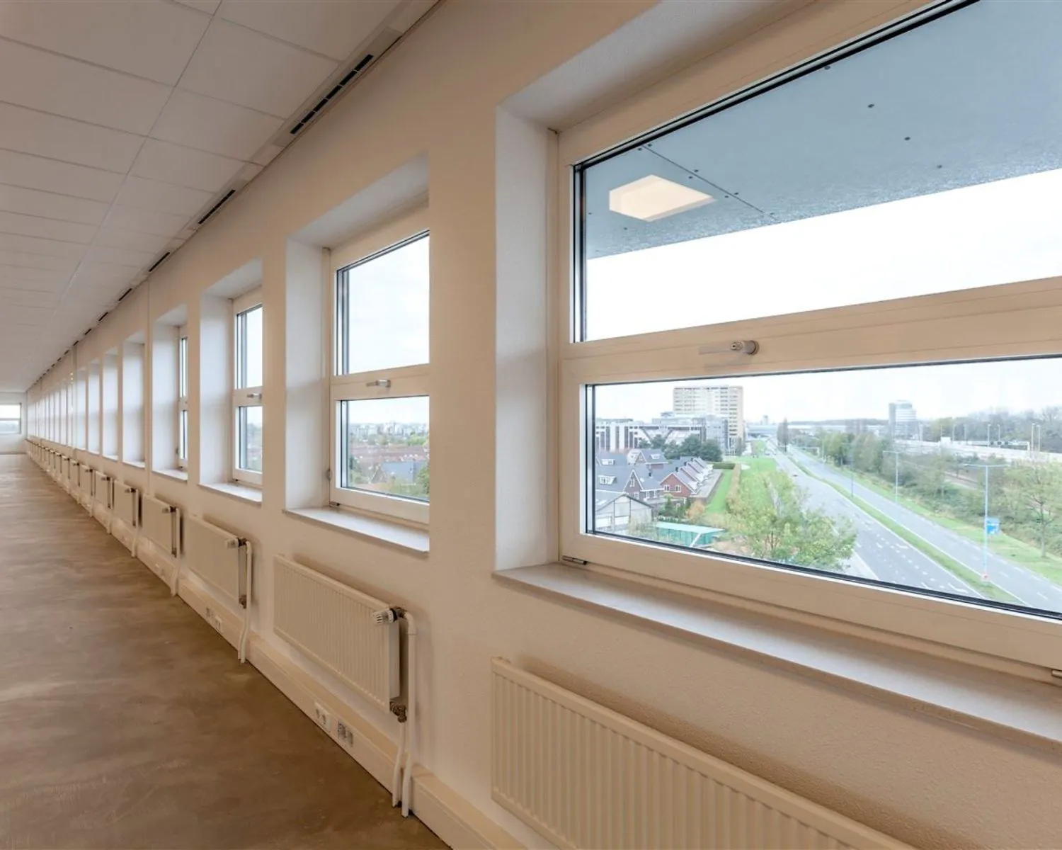 Lange gang in een kantoorruimte aan de Groeneweg 2-5 met grote ramen en uitzicht op een weg en omliggende gebouwen.
