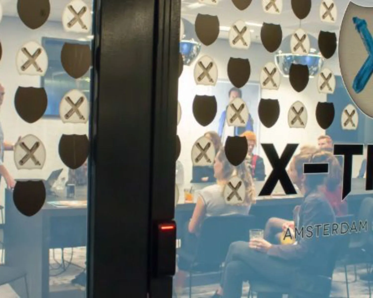 Vergaderruimte met mensen in gesprek, gezien door een glazen deur met X-TEAM Amsterdam-logo erop.