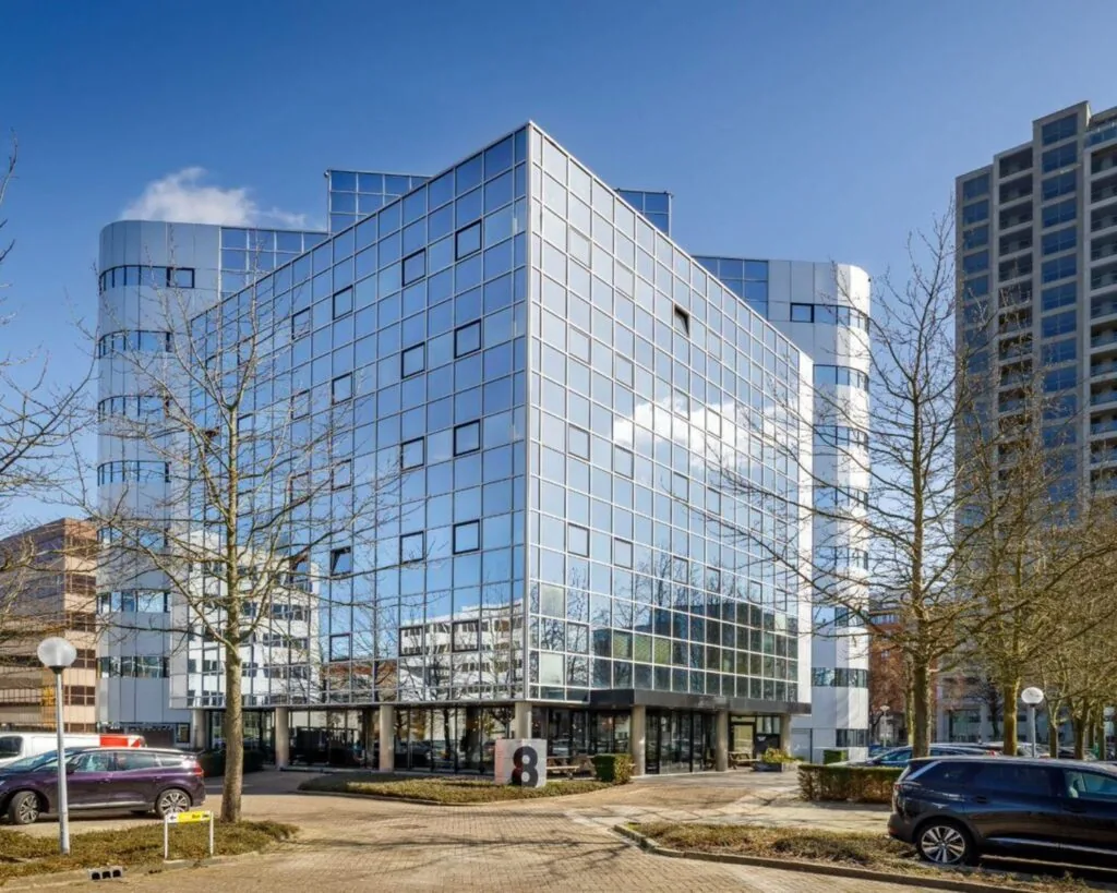 Modern kantoorgebouw met spiegelende gevel aan de Hogehilweg 8 in Amsterdam, omringd door bomen en geparkeerde auto's.