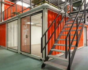 Moderne kantoorruimte in Houten, opgebouwd uit feloranje zeecontainers met metalen trap en glazen deuren.