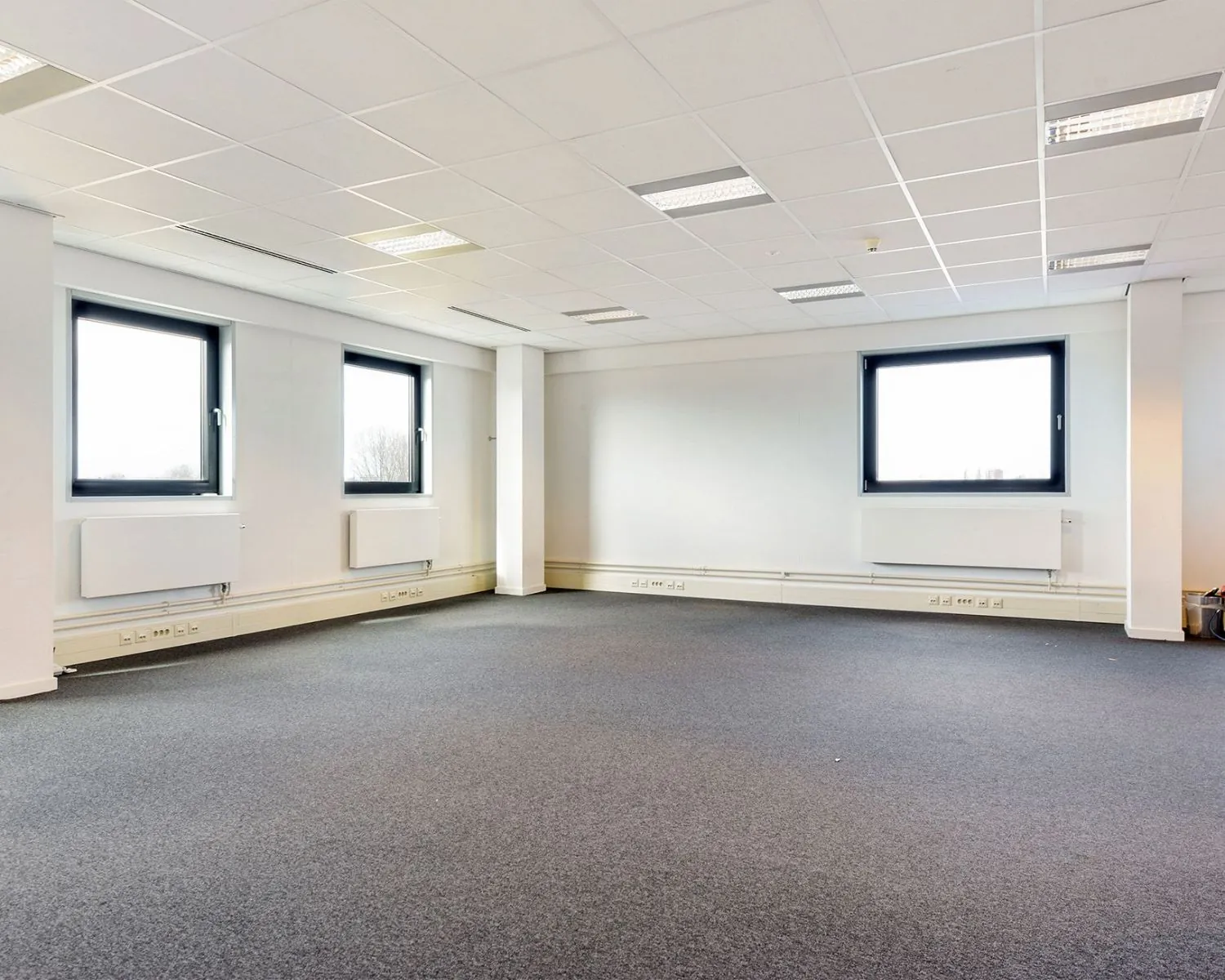 Lichte, lege kantoorruimte met tapijtvloer en drie grote ramen aan de Joan Muyskenweg in Amsterdam.