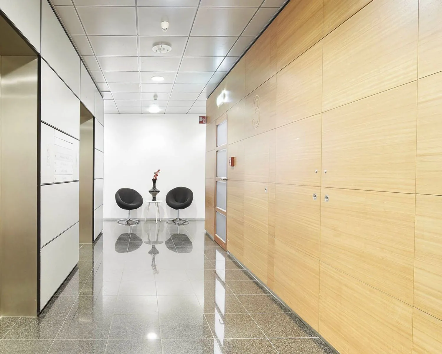 Moderne kantoorhal met glanzende tegelvloer, houten wandpanelen en een zitgedeelte met twee zwarte stoelen aan de Joan Muyskenweg 40-146 in Amsterdam.