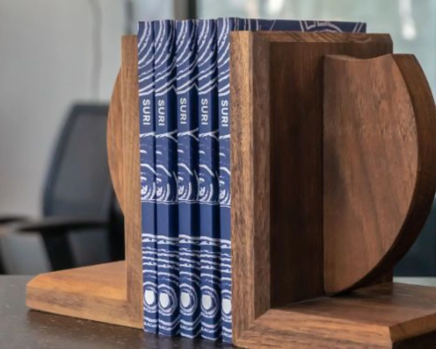 Houten boekenstandaard met vijf blauwe boeken getiteld "SURI" in een moderne kantoorruimte op Prins Bernhardplein 200 in Amsterdam.