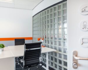 Moderne kantoorruimte met glazen bouwstenen, twee bureaustoelen en een witte tafel met oranje bureaulamp.
