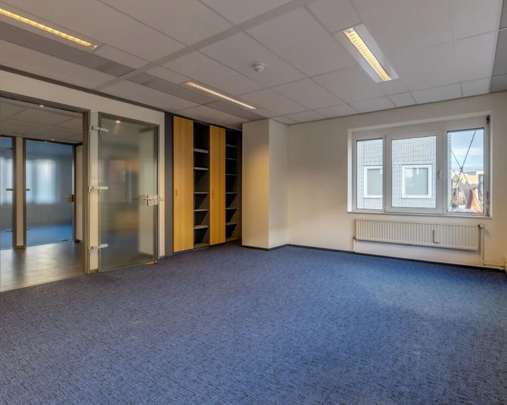 Lichte en nette kantoorruimte met blauw tapijt, ingebouwde kasten en glazen deuren aan de Vossenstraat 6.