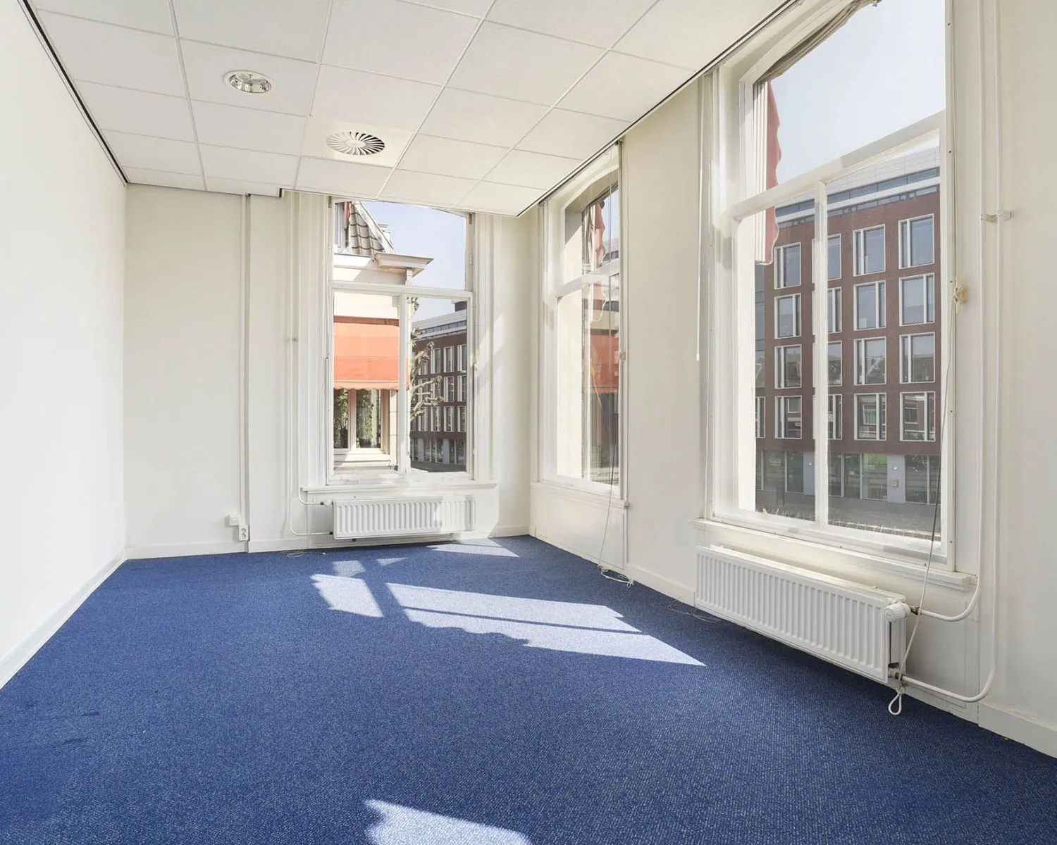 Lichte kantoorruimte met blauwe vloerbedekking en grote ramen aan het Houtplein 22 in Haarlem.