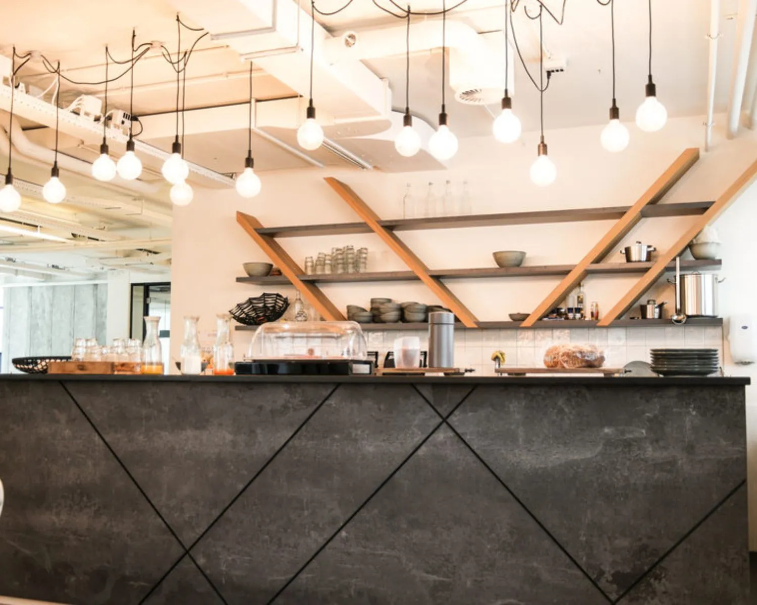 Moderne kantoorkeuken met een donker betonnen barblad, open houten wandplanken en hangende lichtbollen aan het plafond.