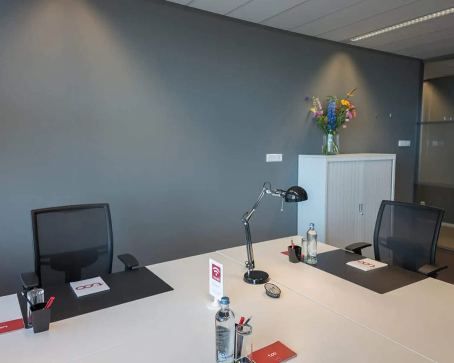 Moderne kantoorruimte aan het Smallepad 32 in Amersfoort met twee werkplekken, een grijze muur en een kast met bloemenvaas.