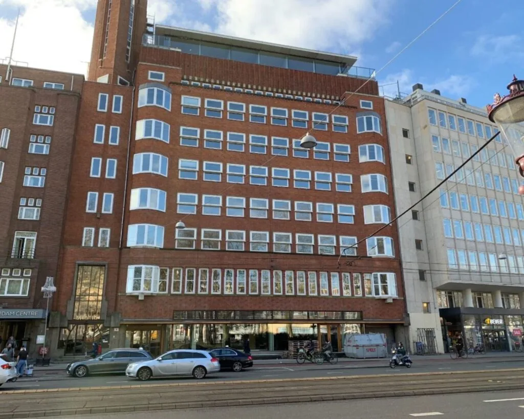 Voorgevel van een roodbruin kantoorgebouw aan de Stadhouderskade 5 in Amsterdam met grote ramen en winkels op de begane grond.