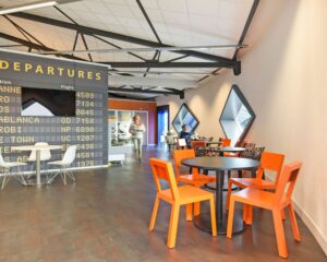 Moderne kantoorruimte in SugarCity Halfweg met feloranje stoelen, ronde tafels en een wand in luchthavenstijl met vluchtnamen.