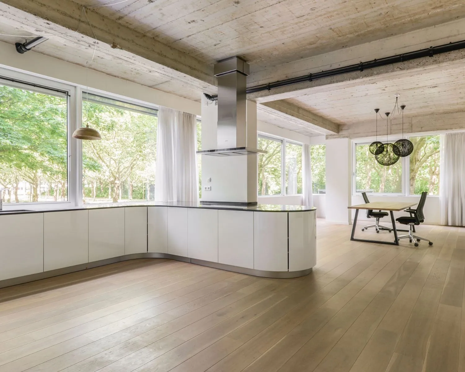 Lichte en moderne kantoorruimte met open keuken, houten vloer en grote ramen met uitzicht op groen.