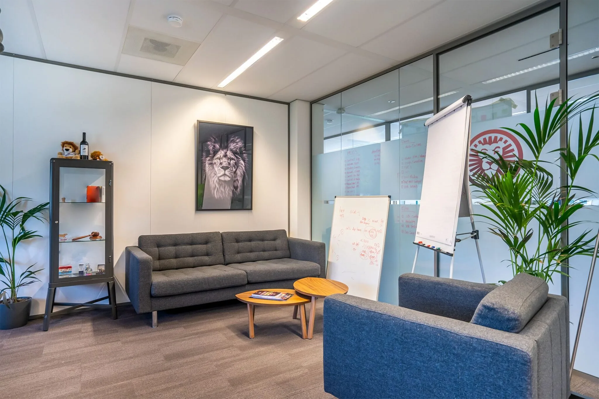 Moderne vergaderruimte met grijze zitbanken, een whiteboard, flip-over en een kunstwerk van een leeuw aan de muur.