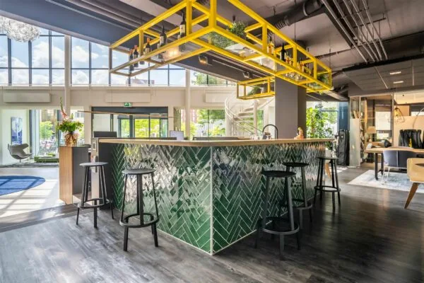 Moderne kantoorruimte met hoge ramen en een centrale bar met groene tegels en gele hangrekken.