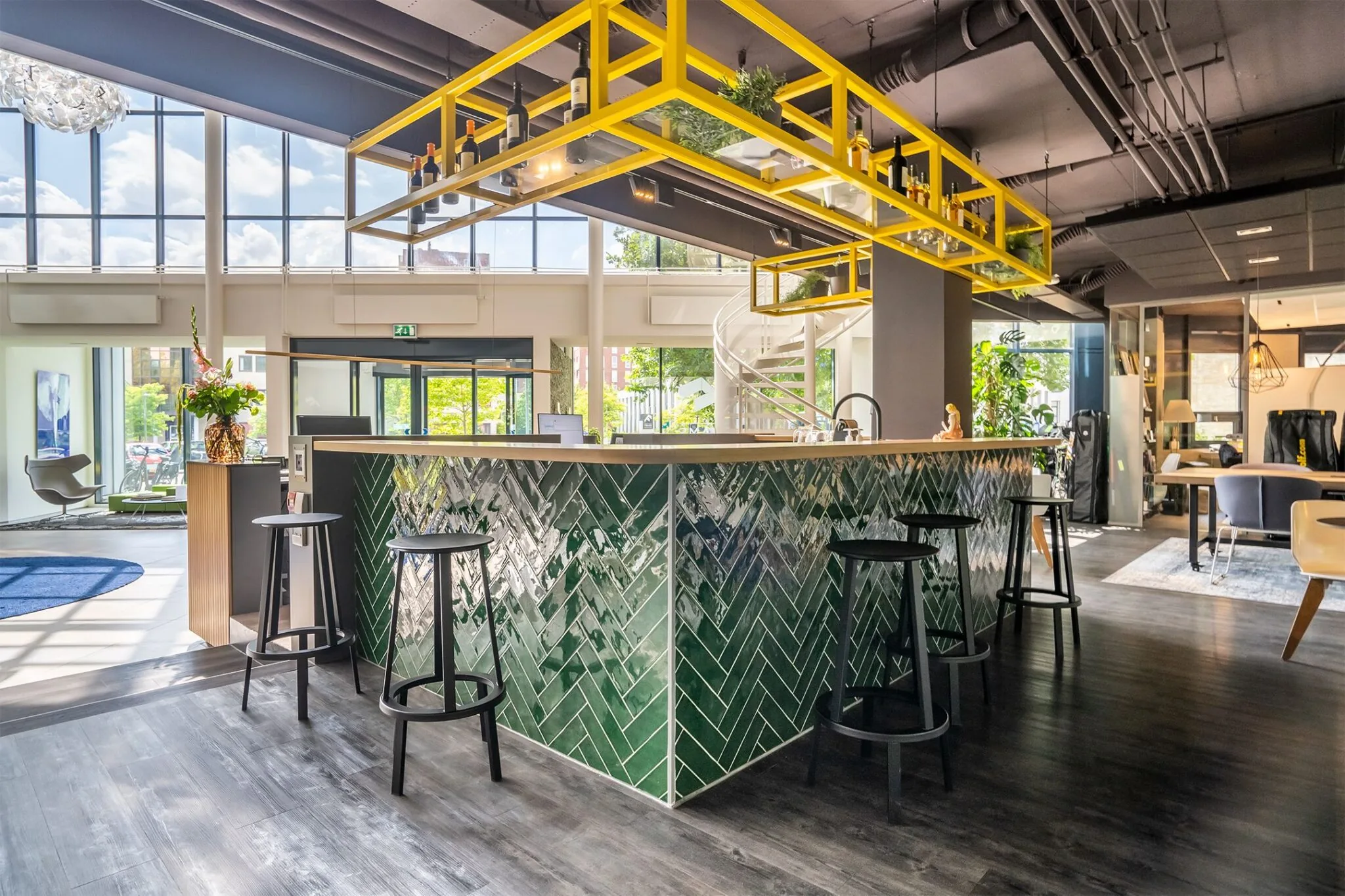 Moderne kantoorruimte met hoge ramen en een centrale bar met groene tegels en gele hangrekken.