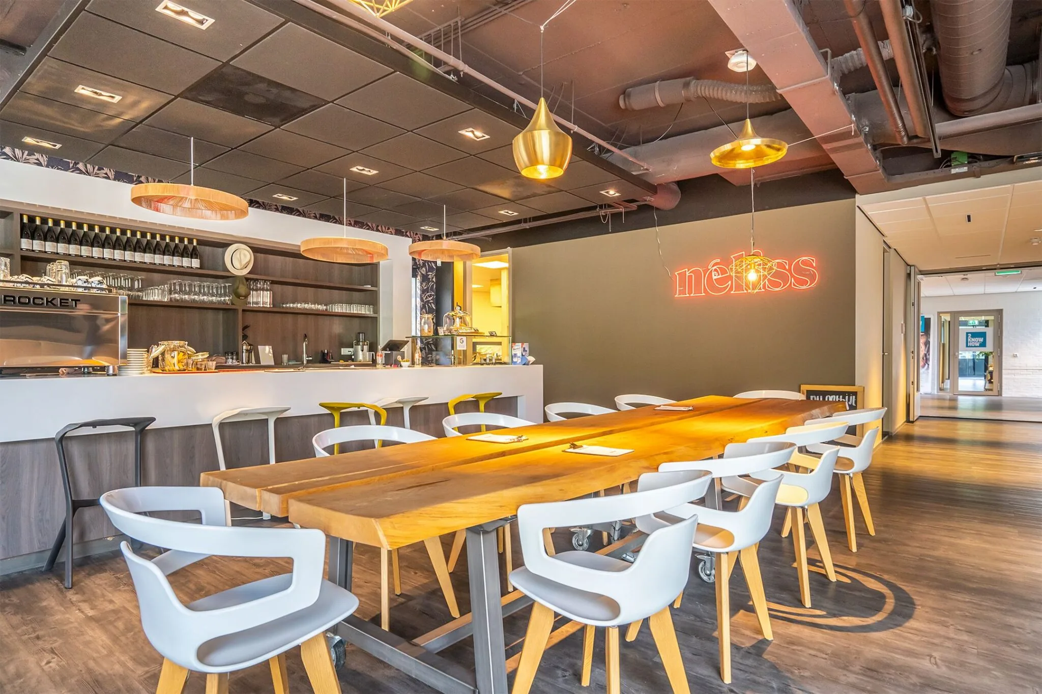 Moderne kantoorkantine met lange houten vergadertafel, open bar en neonverlichting met de tekst "néhss".