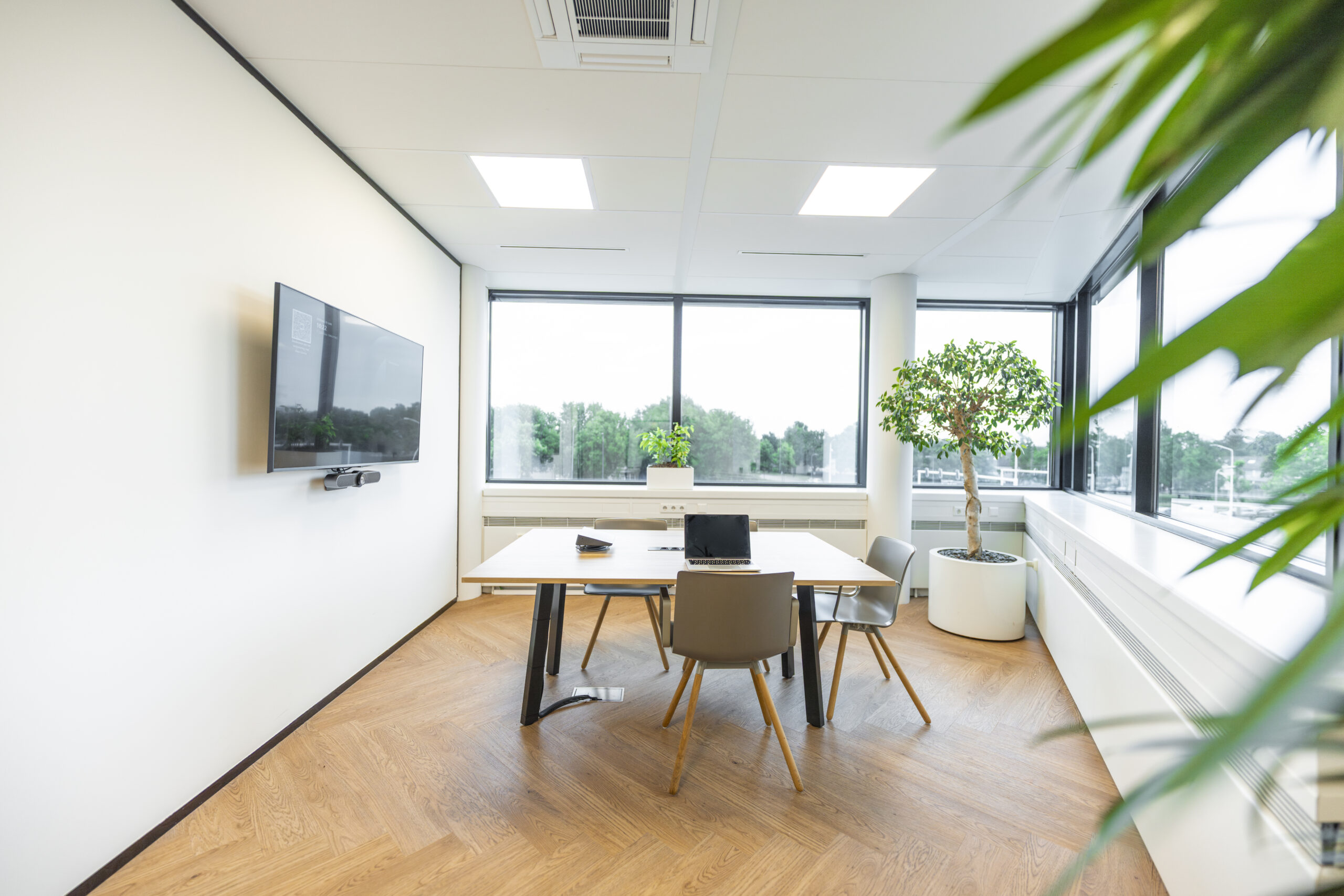 Modern kantoor met vergadertafel, groot beeldscherm en veel natuurlijk licht door grote ramen.