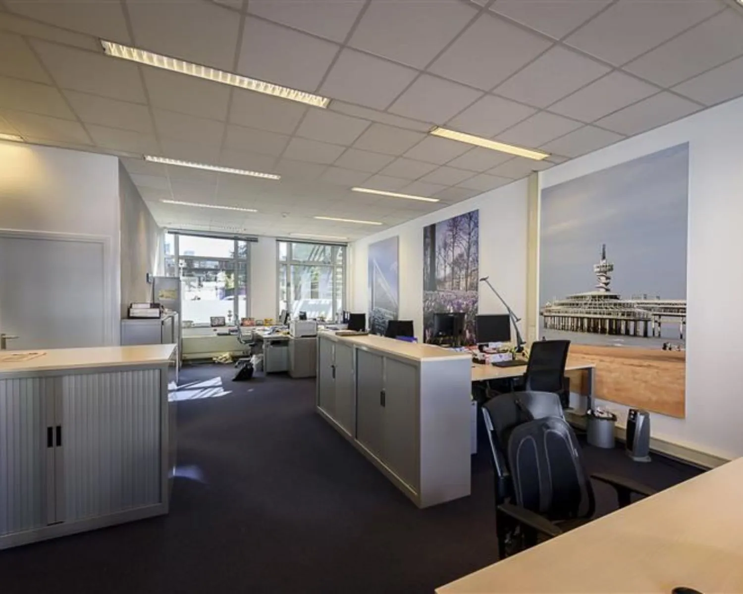 Modern kantoor met meerdere werkplekken, grote fotoposters aan de muur en natuurlijk licht via grote ramen.