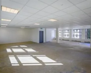 Leeg kantoor met systeemplafond, betonnen vloer en grote ramen aan één zijde.