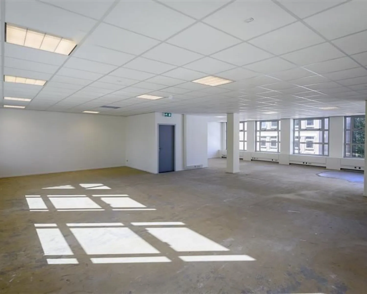 Leeg kantoor met systeemplafond, betonnen vloer en grote ramen aan één zijde.