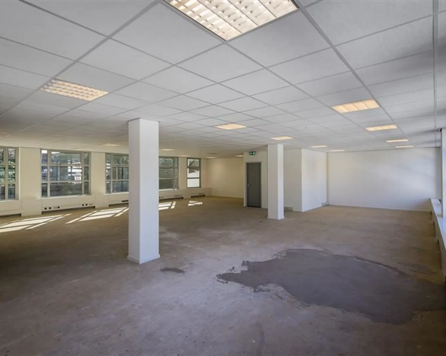 Lege, ruime kantoorruimte met systeemplafond, betonnen vloer en grote ramen aan één zijde.