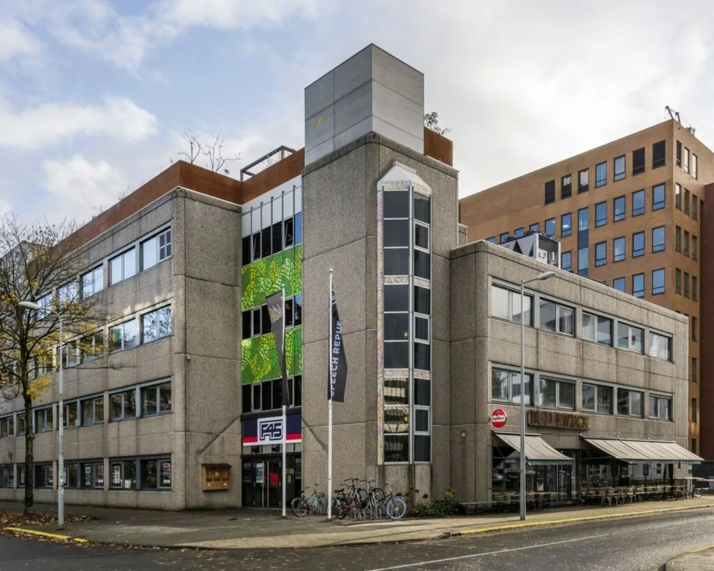 Grijs kantoorgebouw aan de Hogehilweg 20 in Amsterdam met vlaggen, fietsenrekken en horecagelegenheid op de begane grond.
