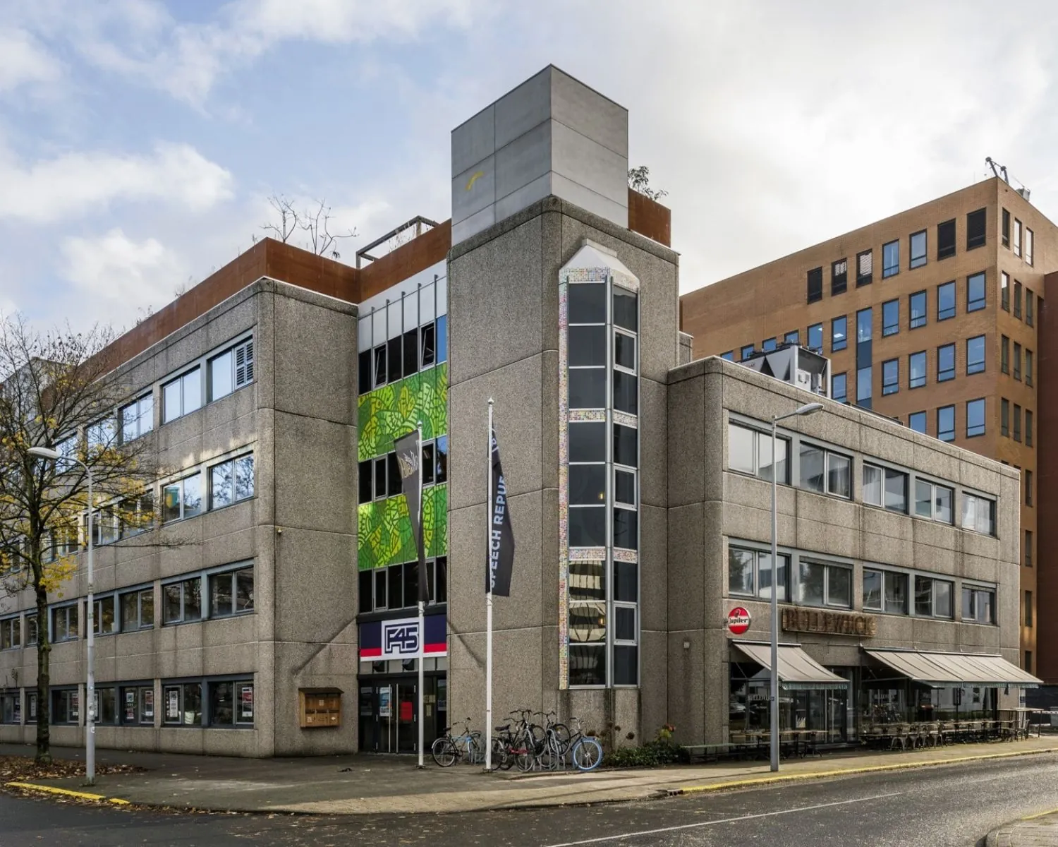 Grijs kantoorgebouw aan de Hogehilweg 20 in Amsterdam met vlaggen, fietsenrekken en horecagelegenheid op de begane grond.