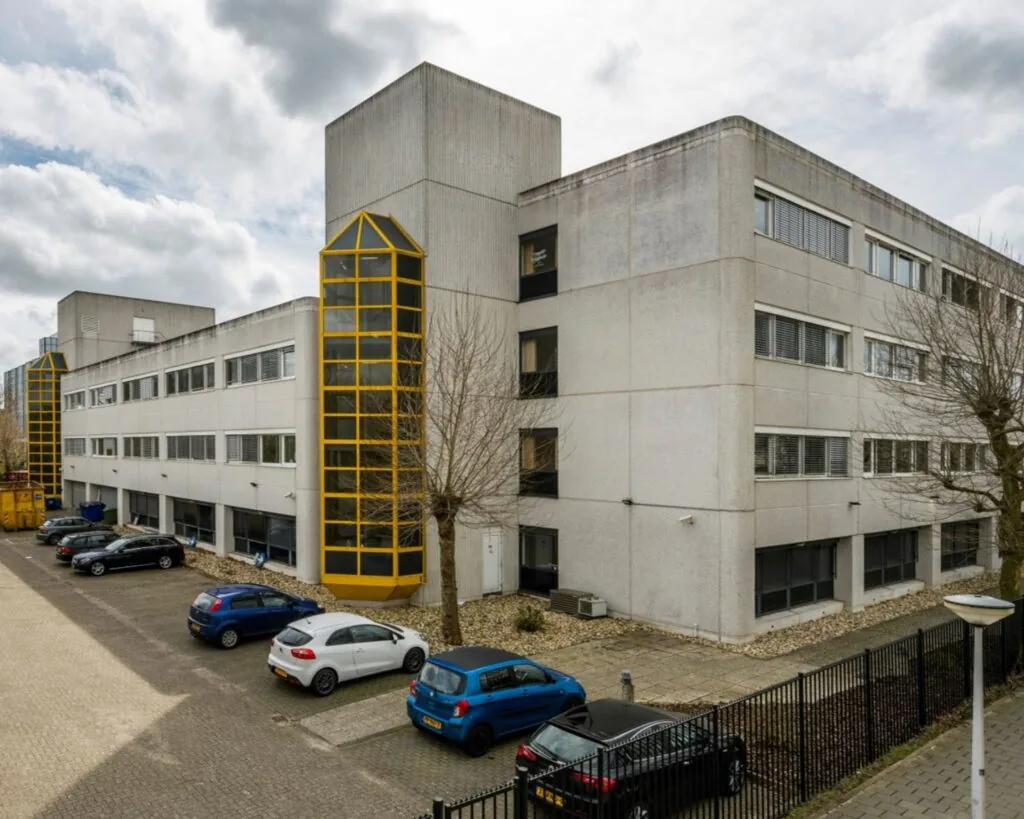 Grijs kantoorgebouw aan de Diemermere 1 in Diemen met gele accenten en een parkeerplaats ervoor.