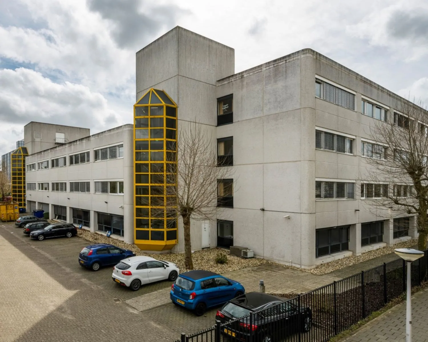 Grijs kantoorgebouw aan de Diemermere 1 in Diemen met gele accenten en een parkeerplaats ervoor.